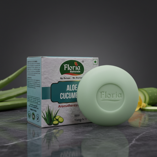 A round pale green Aloe Cucumber bar soap bar in front of its white and teal packaging box, set on a dark reflective surface with aloe vera leaves in the background.